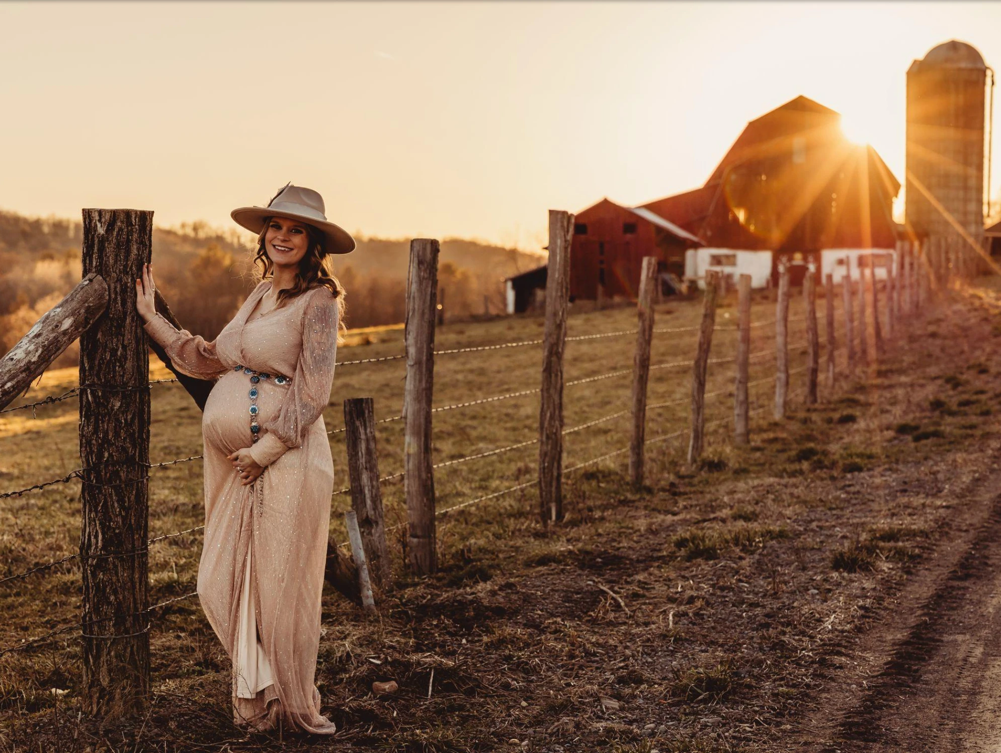 An expectant mother at golden hour, resting a hand on her belly while leaning on a weathered farm fence, a barn and silos lit by the setting sun behind her